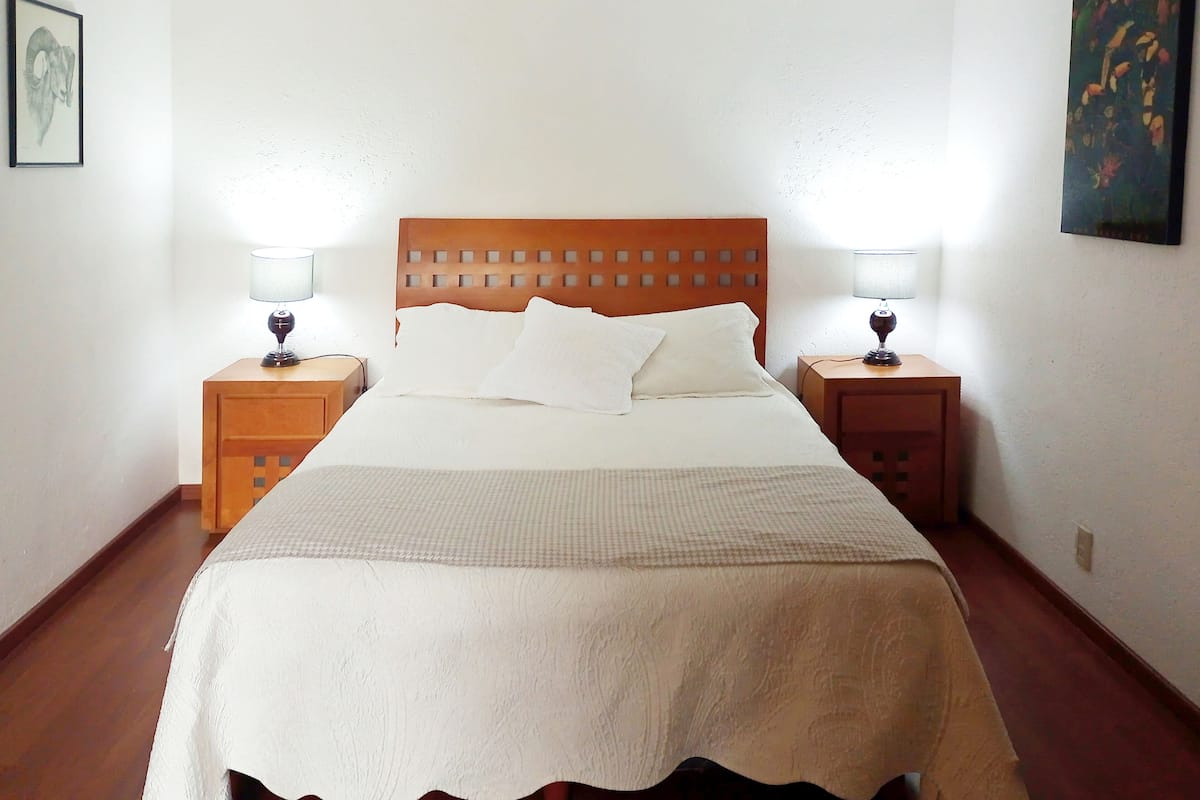 Una cama bien hecha con cabecera de madera, dos mesitas de noche con lámparas y un cuadro en la pared.