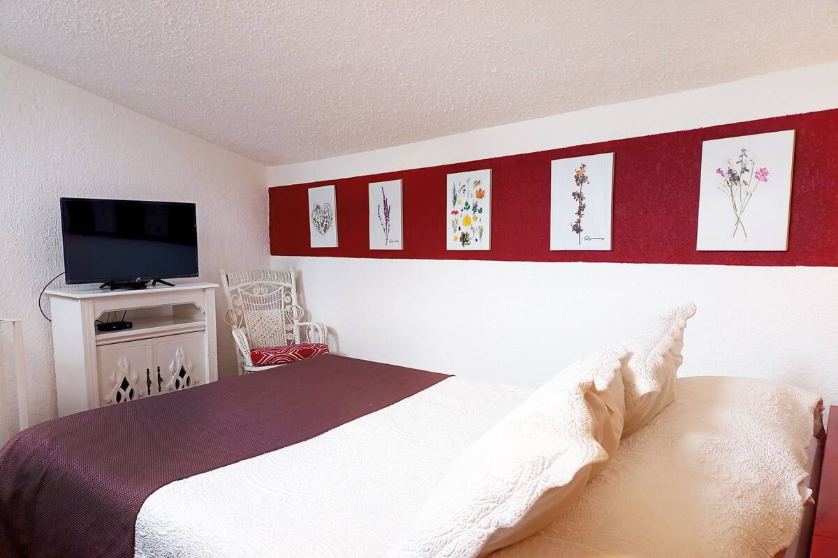 Un dormitorio con cama, televisor sobre un mueble, una silla y una pared decorada con marcos.