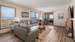 A spacious living room with a grey sofa, a coffee table, and a view of the beach through large windows.