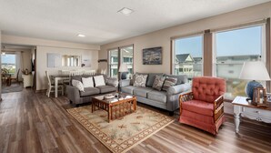 A spacious living room with hardwood flooring, a grey sofa set, a red armchair, and a dining area with a white table and chairs.