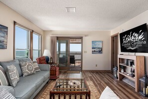 A living room with a sofa, a coffee table, a television, and a bookshelf. - Carolina Ocean Views: Dog Friendly, Ocean Views (Kill Devil Hills)
