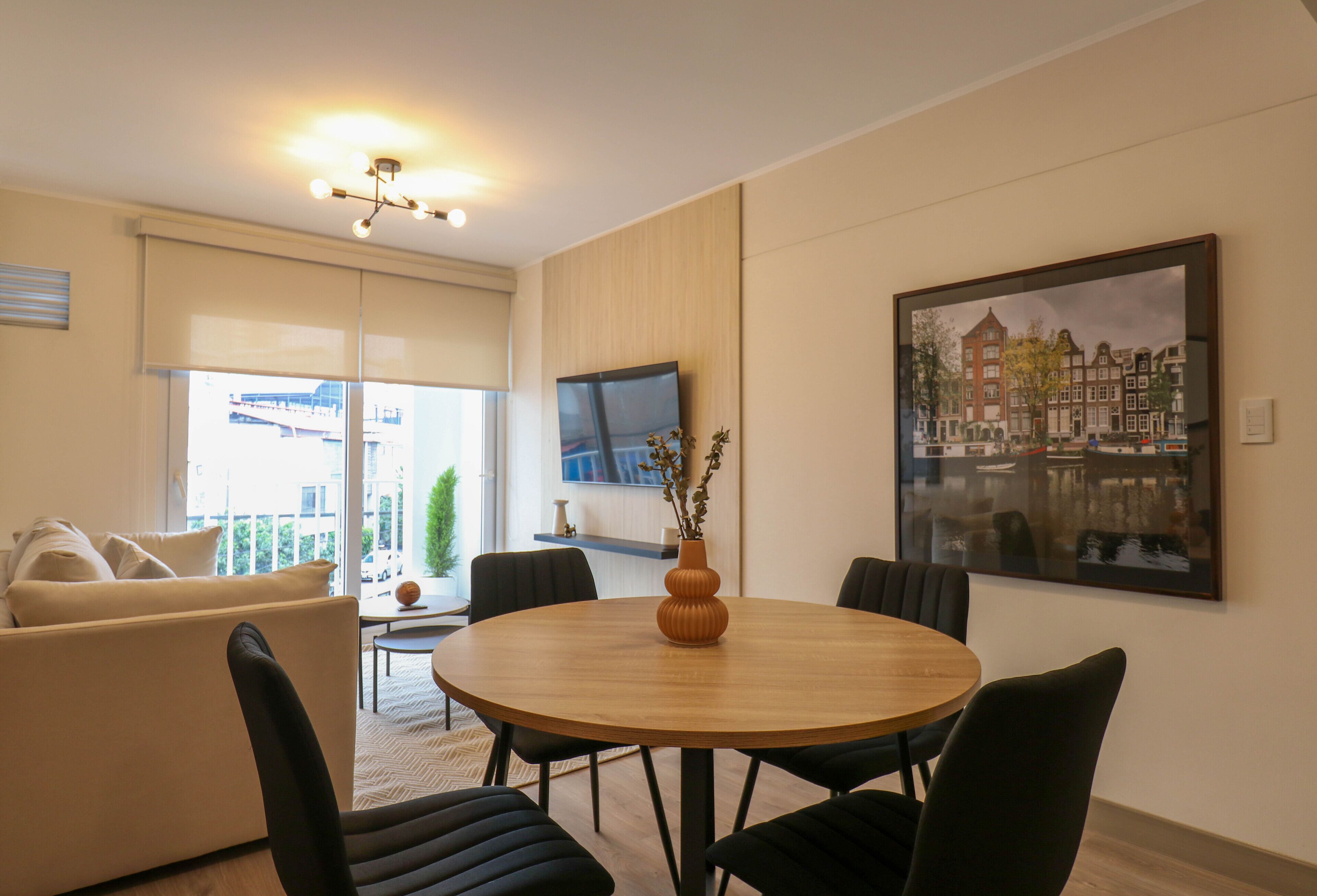 A modern living room with a round wooden table, black chairs, a beige sofa, a flat-screen TV mounted on the wall, and a large framed painting of a cityscape.