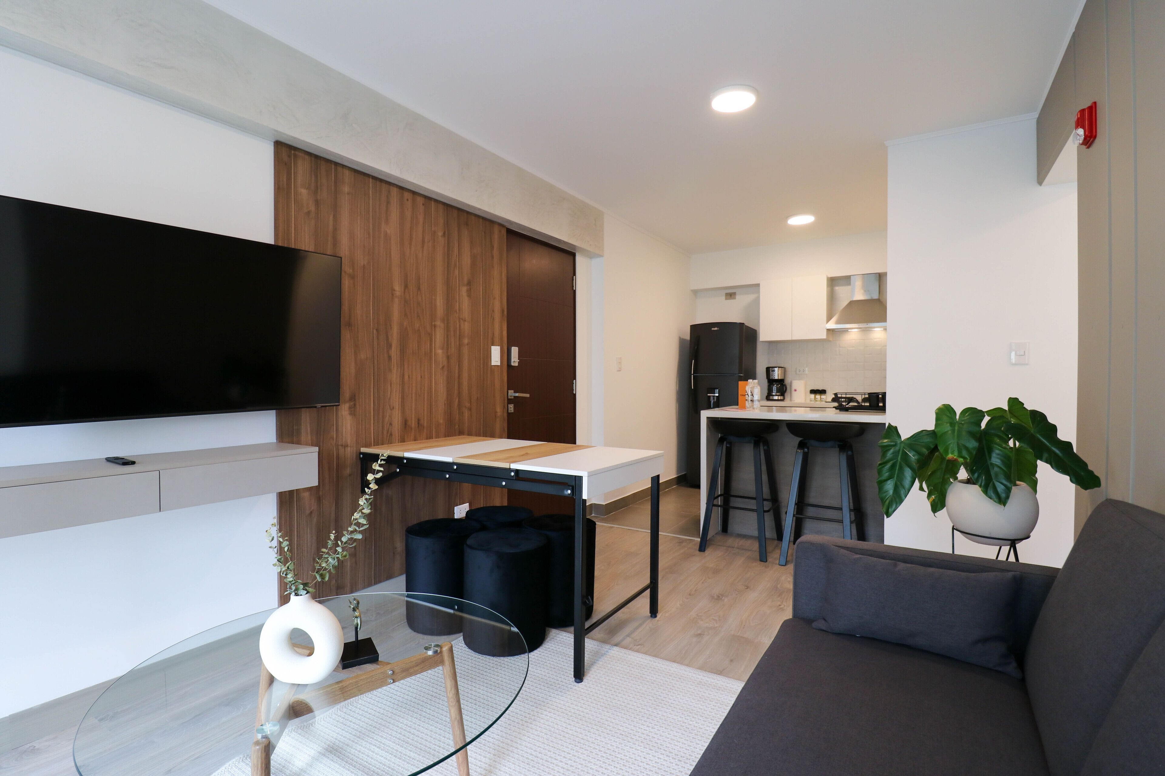 A modern living room with a flat-screen TV, a sofa, a coffee table, and a kitchen area in the background.
