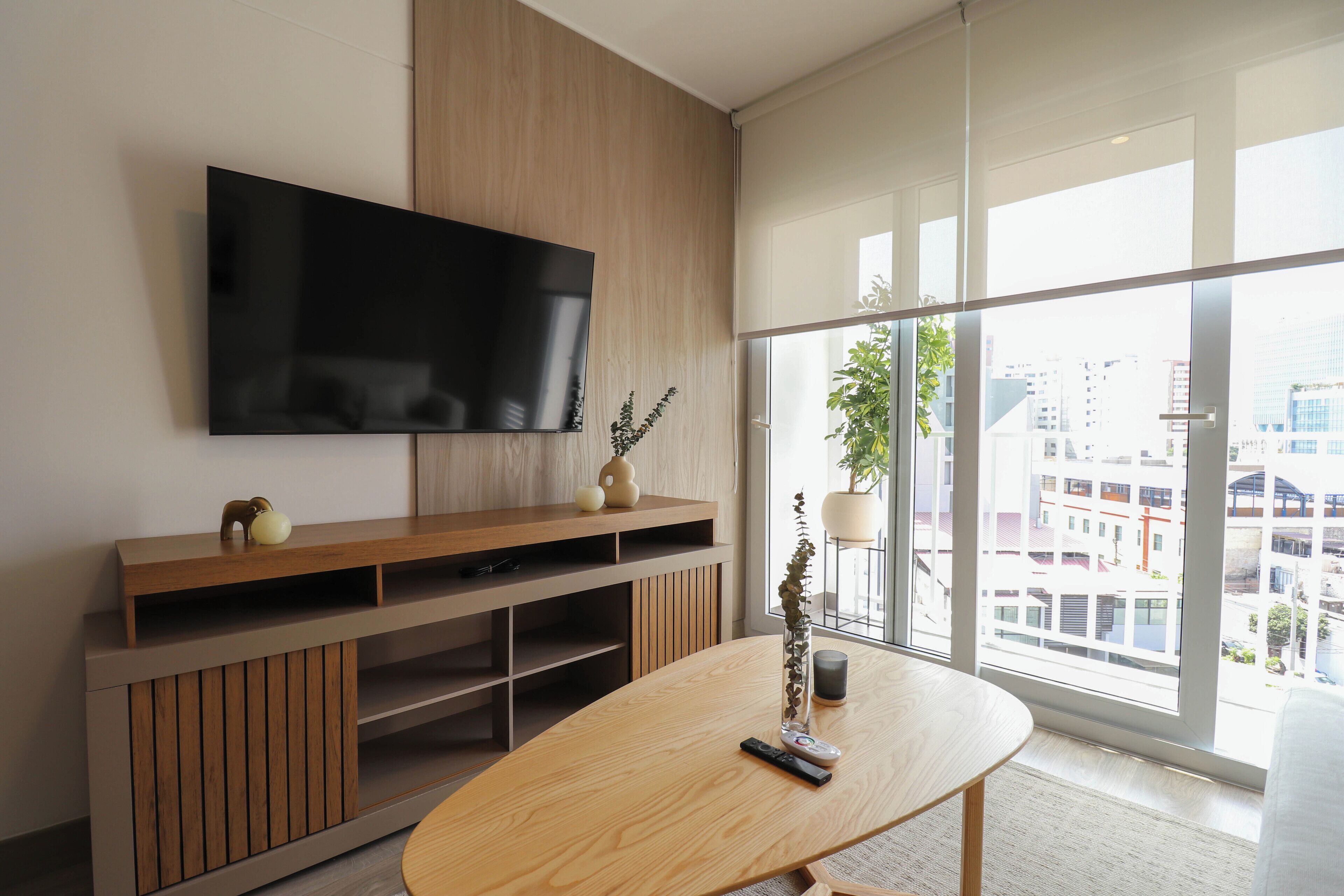 A modern living room with a large flat-screen TV mounted on a wooden wall, a wooden entertainment unit below, and a large window offering a city view.