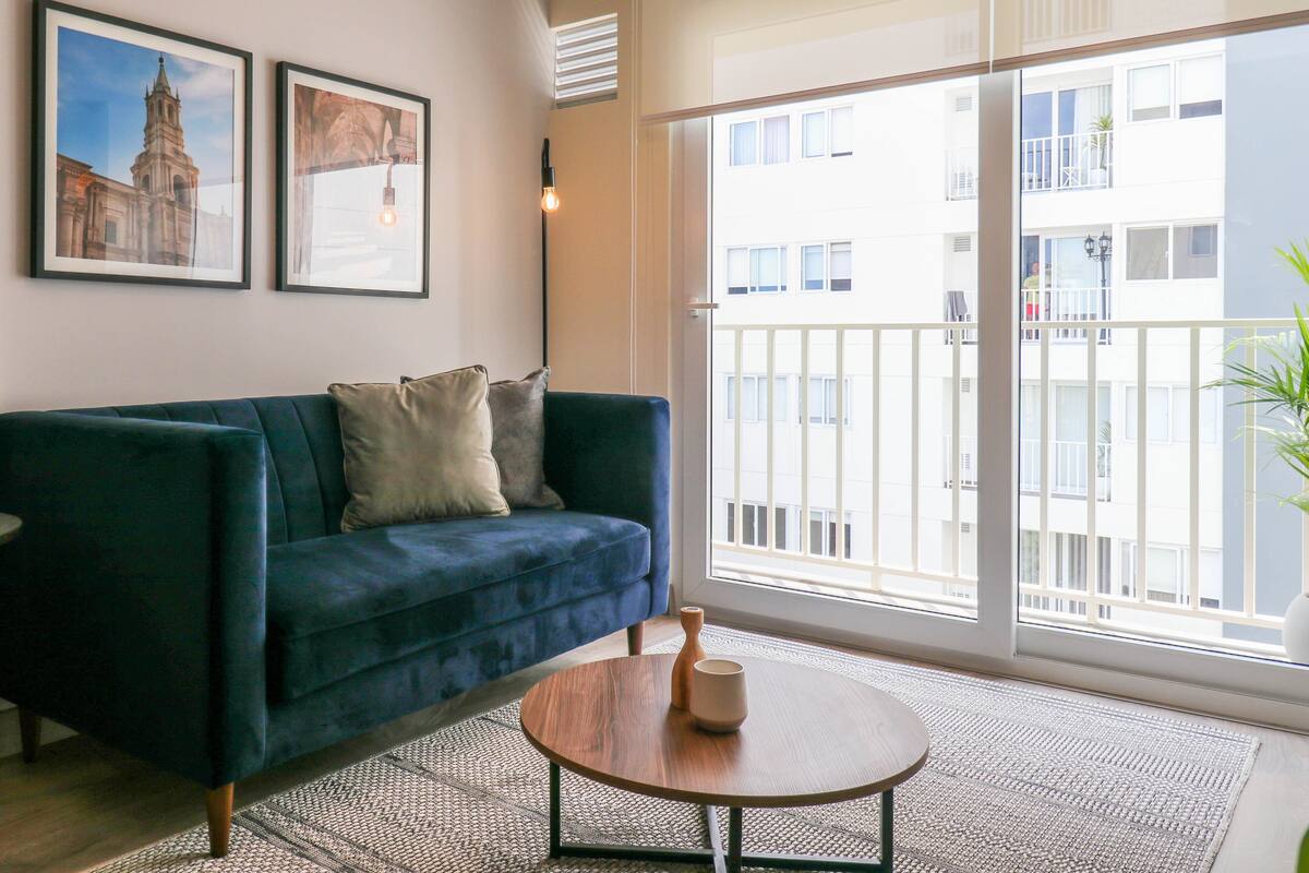 A modern living room with a dark blue sofa, two framed pictures on the wall, a round wooden coffee table, and a large sliding glass door leading to a balcony with a view of a building.