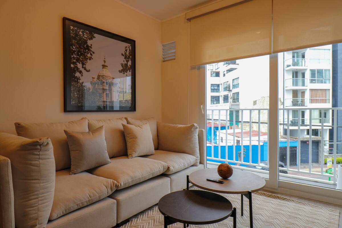 A modern living room with a beige sofa, a small round table, and a large window offering a view of a cityscape.
