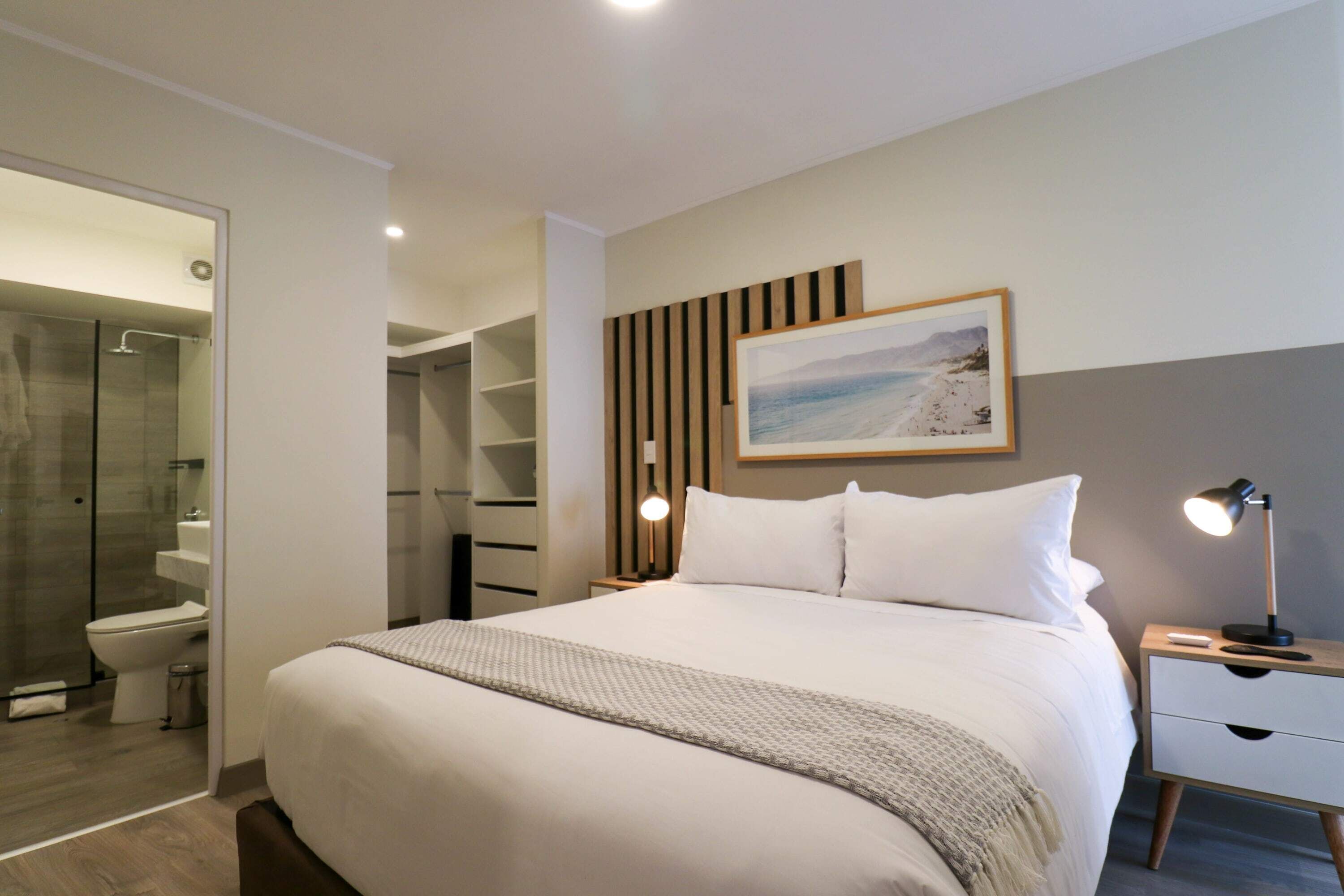 A modern hotel room with a large bed, a bedside table with a lamp, a framed picture on the wall, and a walk-in closet.
