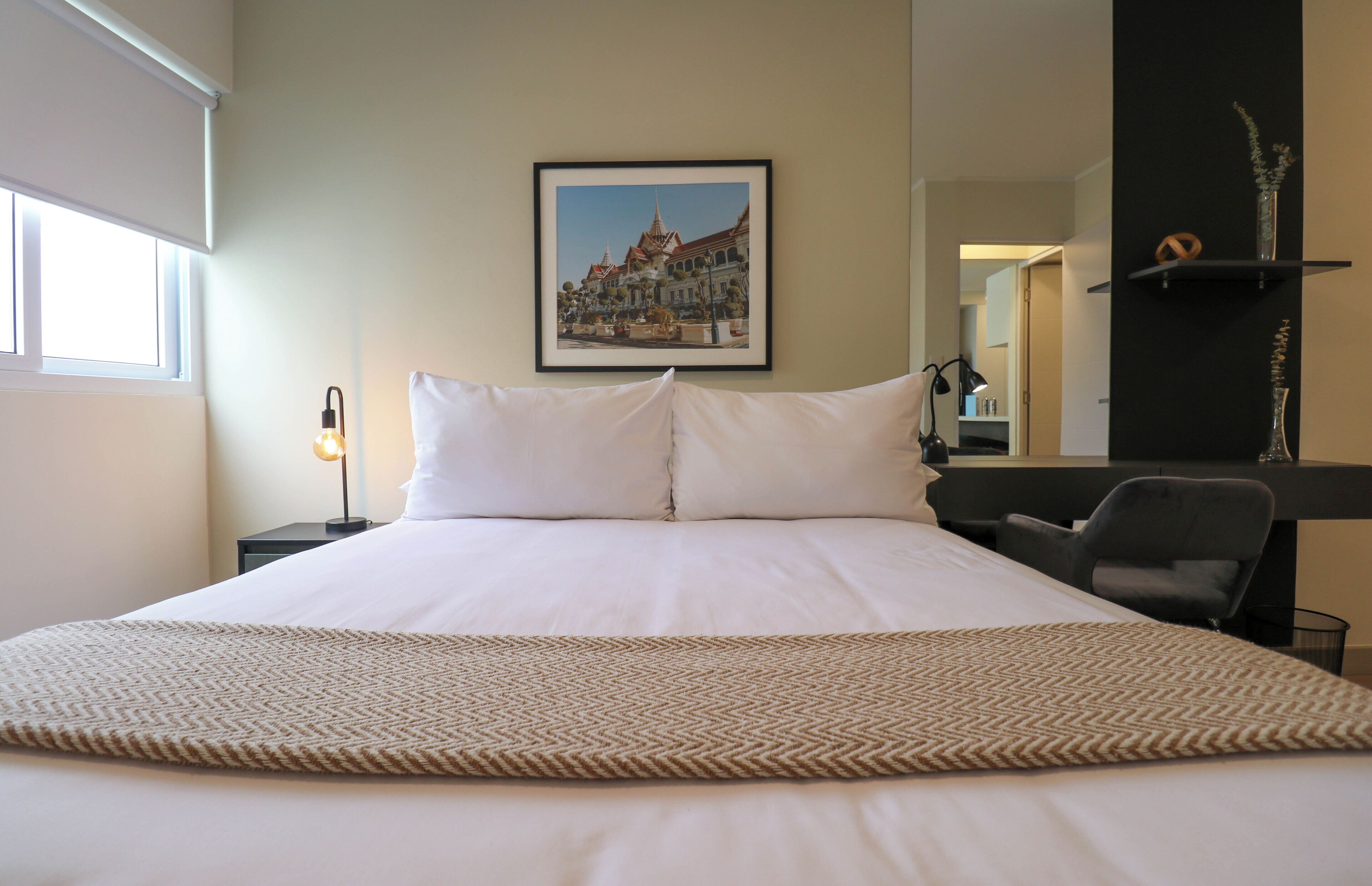A neatly made bed with white linens, a framed picture of a cityscape on the wall, a desk with a chair, and a lamp.