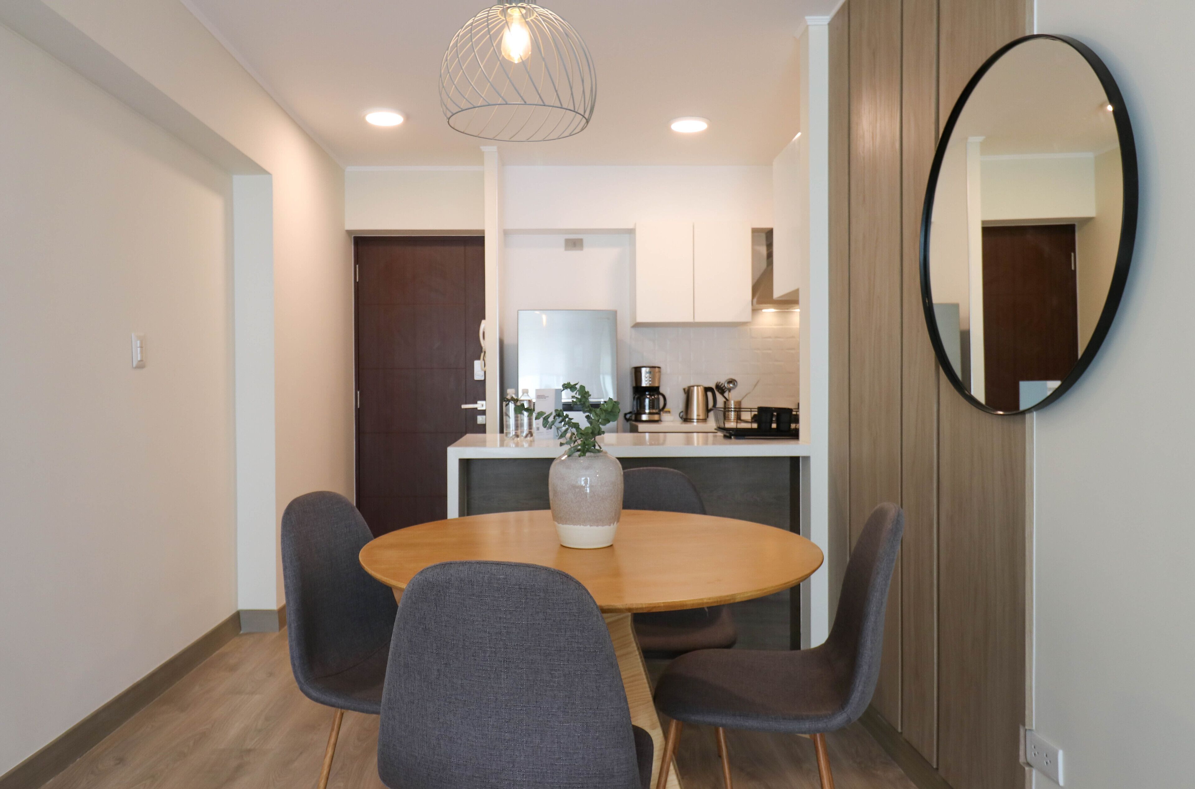 Un coin repas avec une table ronde, quatre chaises grises, un vase avec une plante et un grand miroir au mur.