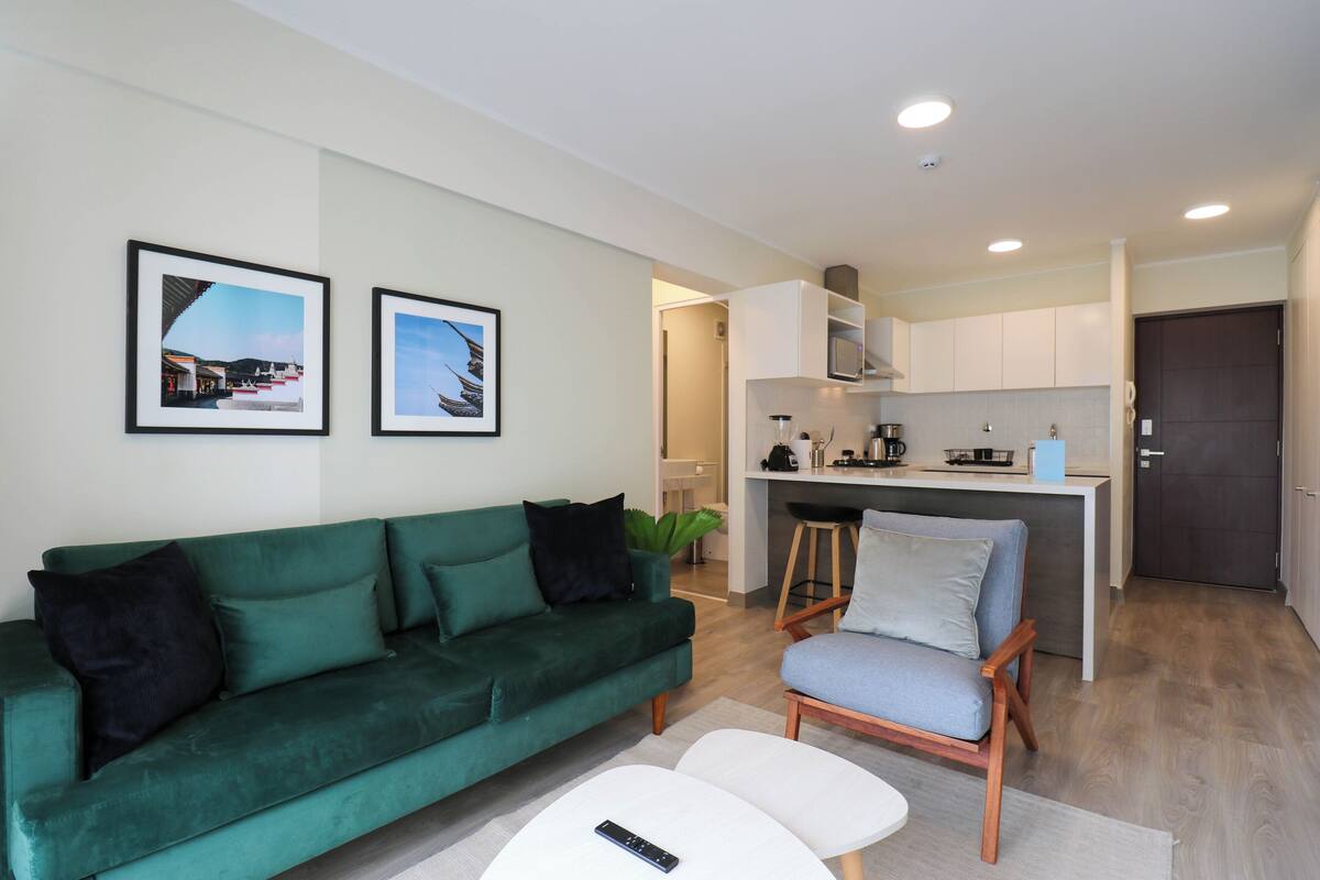 A modern living room with a green sofa, a small white table, and a kitchen area in the background.