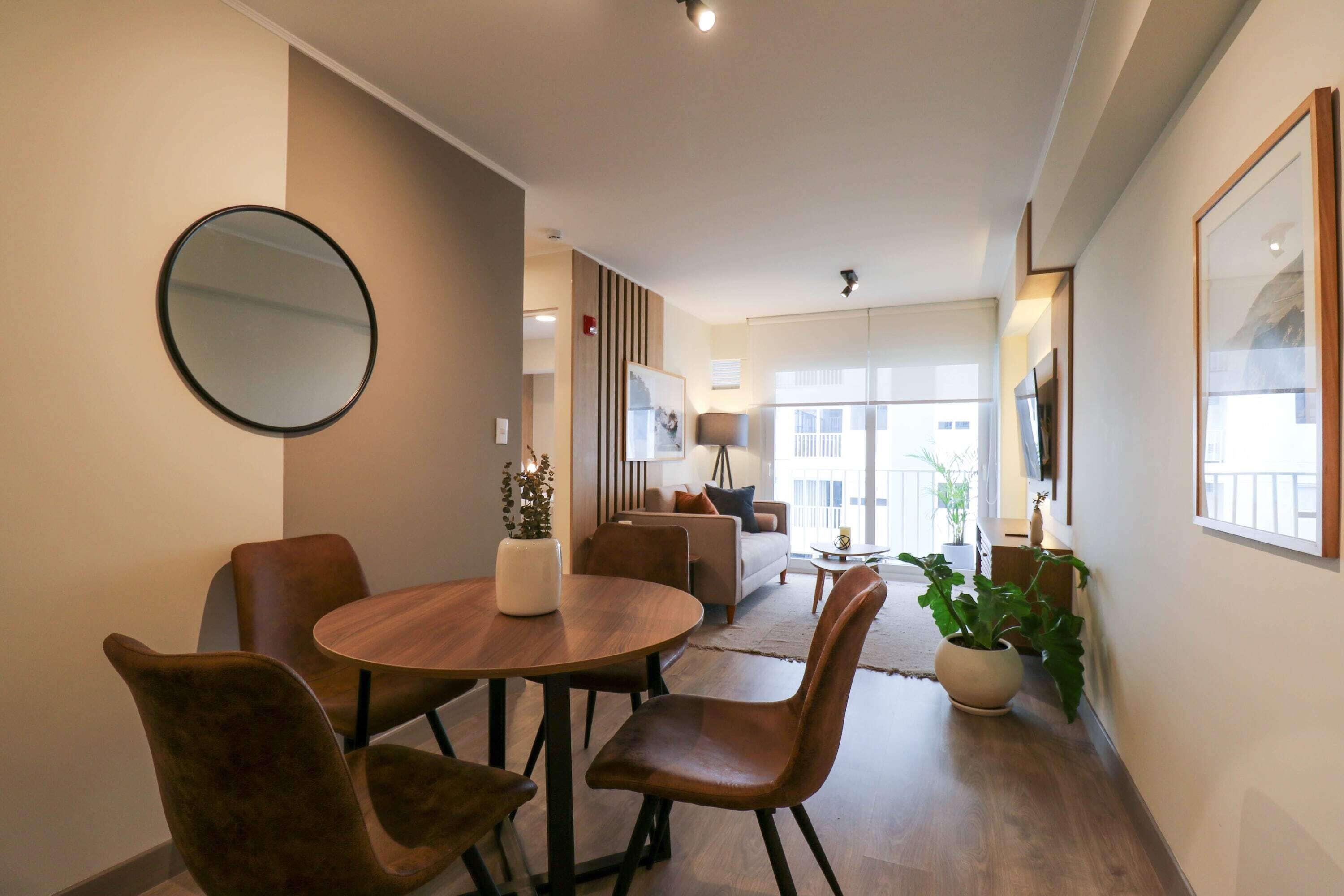 A modern living room with a wooden dining table, brown leather chairs, a round mirror, and a sofa with a view to the outside.