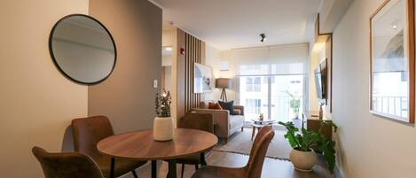 A modern living room with a wooden dining table, brown leather chairs, a round mirror, and a sofa with a view to the outside.