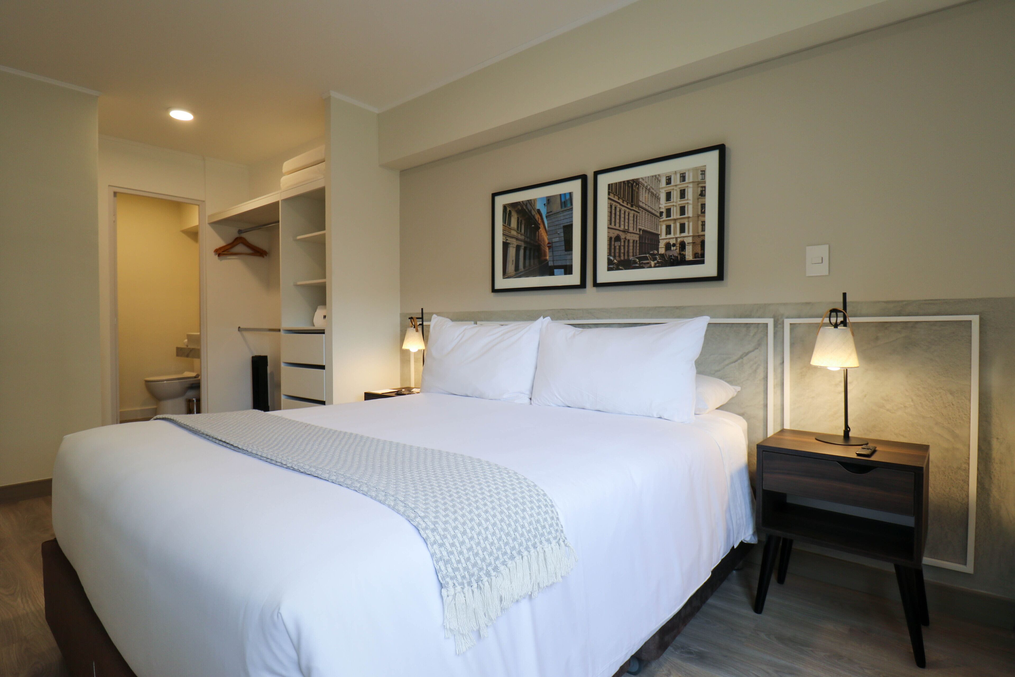 A neatly made bed with white linens and a blue throw, a bedside table with a lamp, and two framed pictures on the wall.
