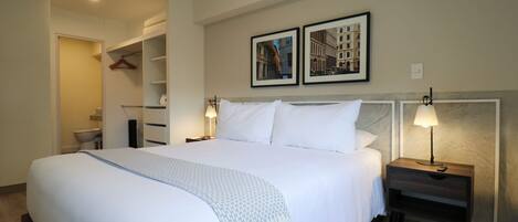 A neatly made bed with white linens and a blue throw, a bedside table with a lamp, and two framed pictures on the wall.