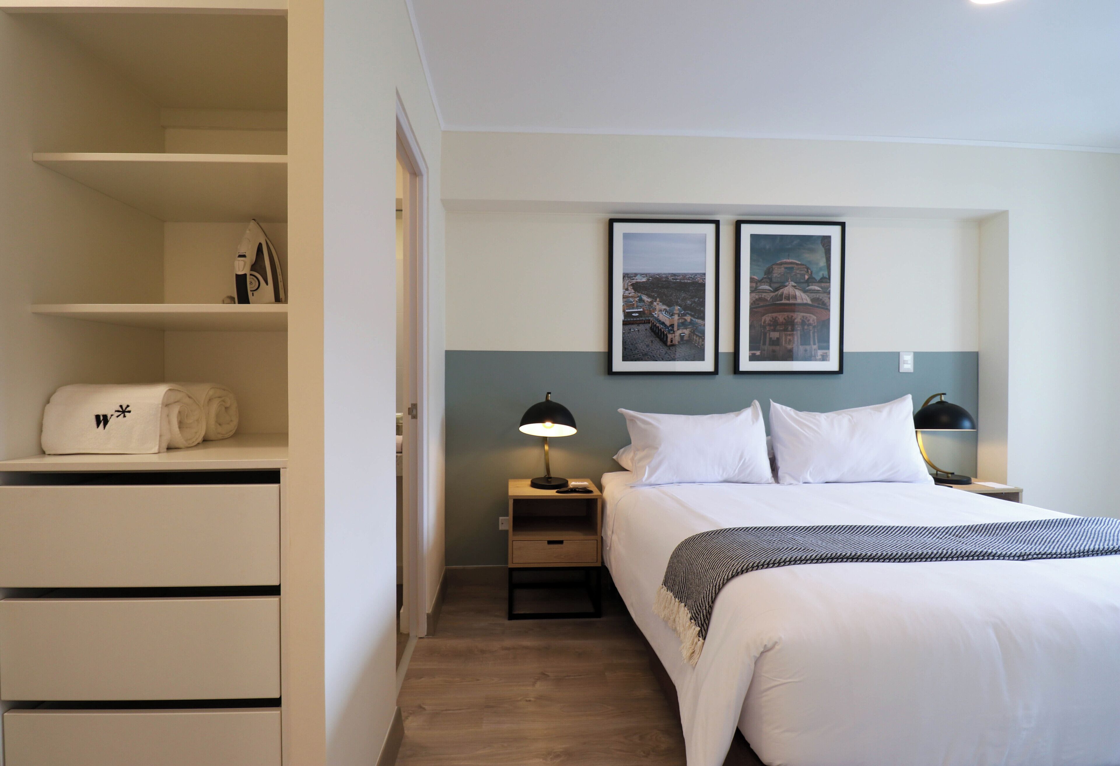 Une chambre d’hôtel avec un lit soigneusement fait, une lampe de chevet, une table de chevet, une étagère avec des serviettes pliées et deux tableaux encadrés au mur.