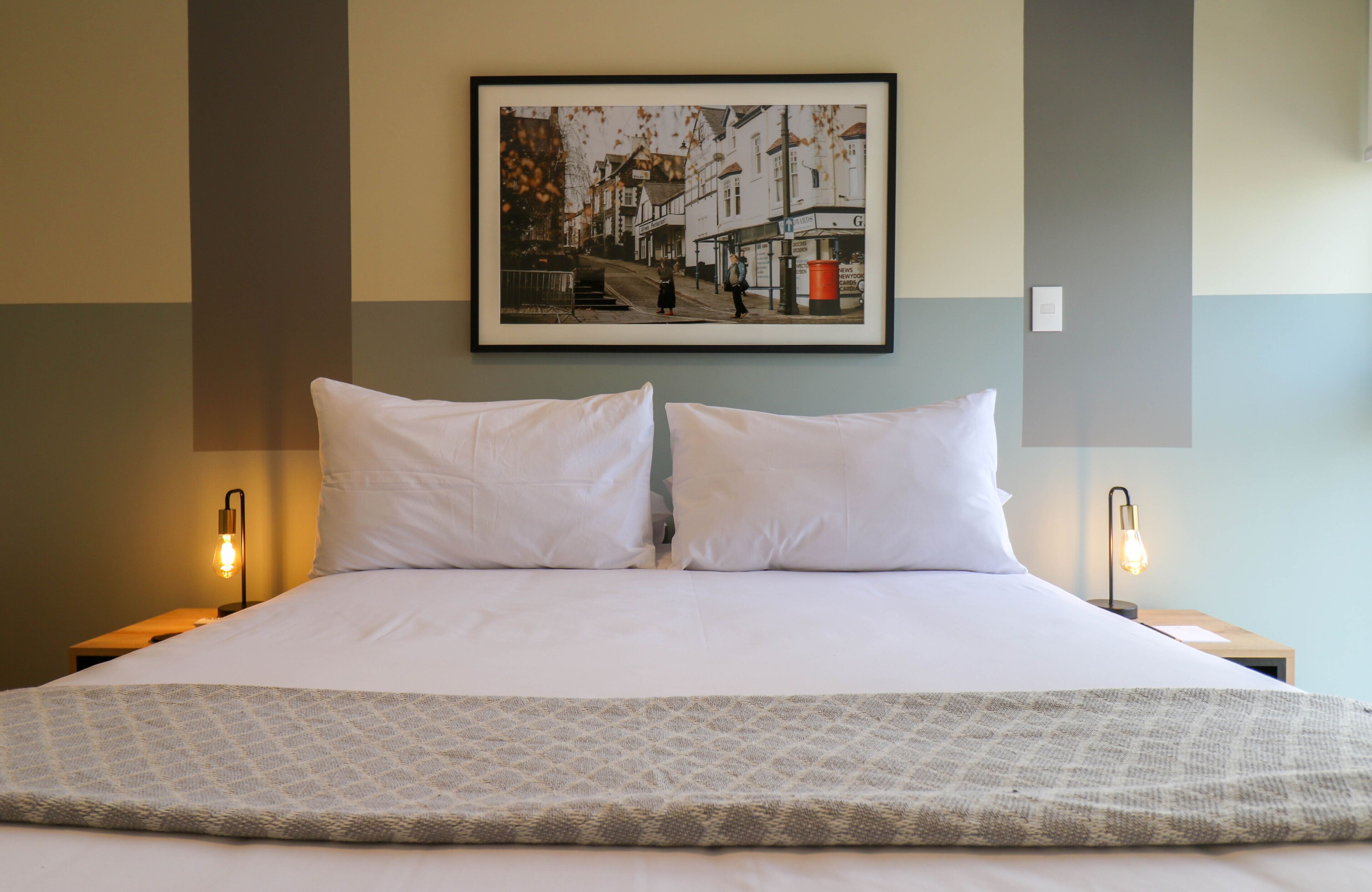 A neatly made bed with white linens and a patterned throw, two bedside lamps, and a framed photograph on the wall.