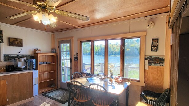 A dining area with a wooden table and chairs, a ceiling fan, and large windows overlooking a body of water. - Relaxing rustic cabin on the lake 25 feet from the shore. (Baileyville)