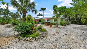 Una casa rodeada de follaje tropical, un patio con mobiliario exterior y un camino de entrada de grava.