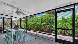 Una sala de estar con ventanales, un ventilador de techo, una mesa de comedor con sillas y vistas al exterior con mucha vegetación.