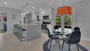 Una cocina moderna con una isla central, taburetes de bar y un comedor con mesa de vidrio y sillas.