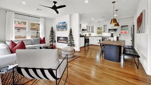 A modern living room with a sofa, a coffee table, and a fireplace. There is a dining area with a table and chairs. The kitchen is visible in the background with white cabinets and a large island.
