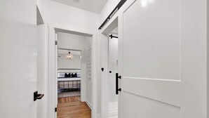 A hallway with white walls and wooden flooring leading to a kitchen area with a countertop and hanging lights.
