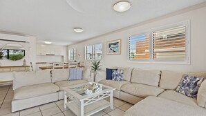 Un salón amplio con un sofá seccional beige, una mesa de centro de vidrio y una zona de cocina con armarios blancos y una mesa de comedor.