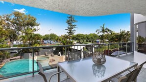 Un balcón con una mesa y sillas de vidrio, con vista a una piscina y exuberante vegetación.