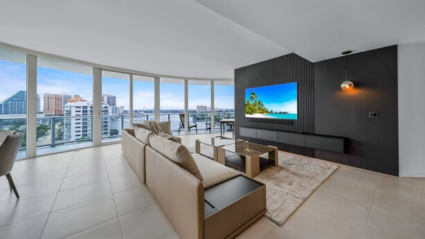 A modern living room with a large TV, comfortable seating, and a city view. - Ocean view Amazing 3BR at Tiffany House (Fort Lauderdale)