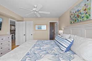 A bedroom with a bed, a ceiling fan, a painting, and a view into the adjacent room.