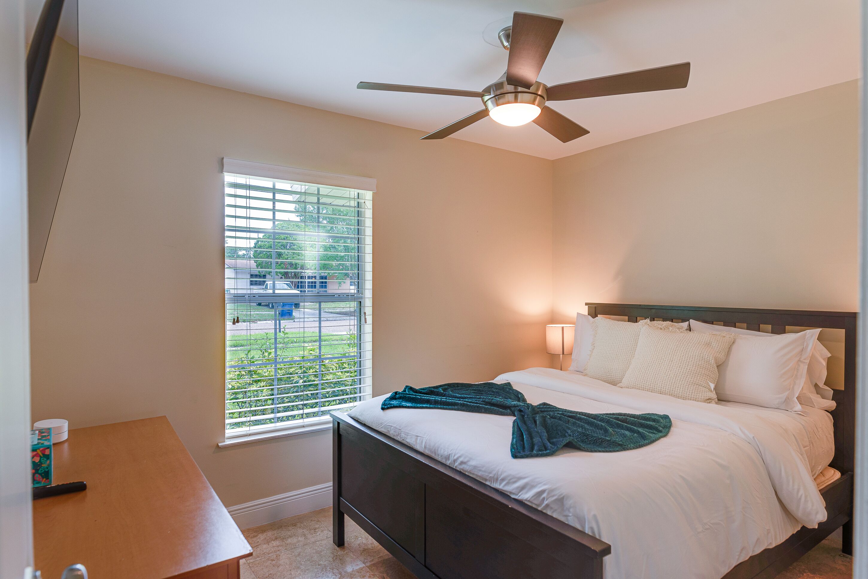 A bedroom with a bed, a ceiling fan, a window with blinds, and a bedside table.