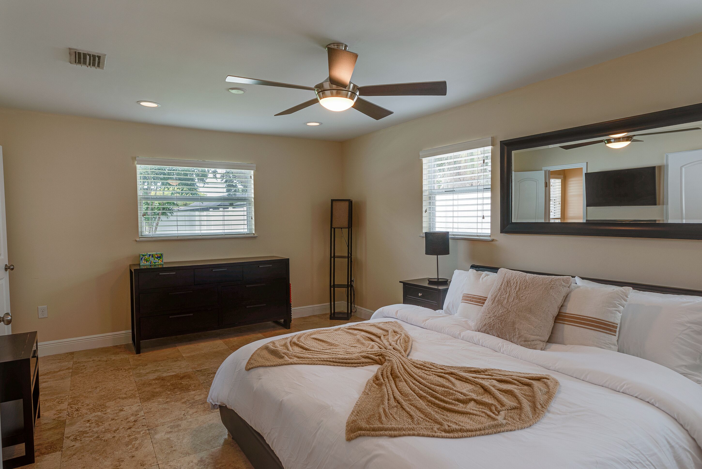 A bedroom with a bed, a ceiling fan, a dresser, a nightstand, a mirror, and a window with blinds.