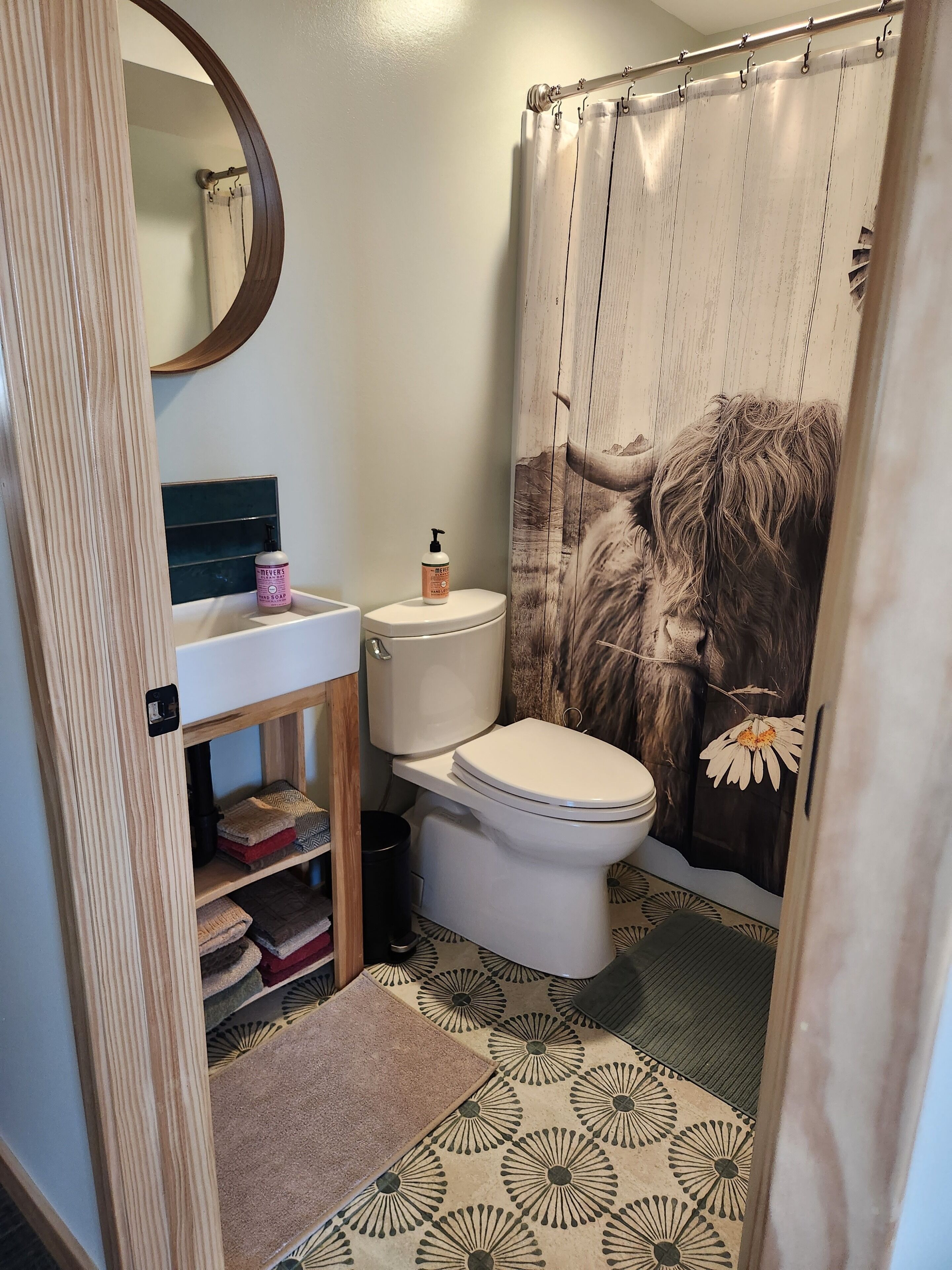 Une salle de bain comprenant des toilettes, un lavabo avec un miroir, un rideau de douche à motifs floraux et des carreaux de sol décorés.