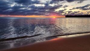 Una playa con un atardecer sobre el agua, nubes en el cielo y una costa distante.