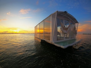 Une structure flottante dotée d’une enceinte transparente, d’un hamac et offrant une vue sur le coucher de soleil sur l’eau.