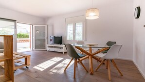 A modern living room with a glass dining table, wooden chairs, a TV on a white cabinet, and a large window allowing natural light. - Vacay L’Escala - Casa Los Pinos, pool and tranquility (l'Escala)