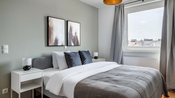 A modern bedroom with a bed, two bedside tables, a nightstand, a window with curtains, and two wall-mounted artworks. - Lovely Mariahilf 2BR w/ Elevator, nr Shops & Cafes, by Blueground (Vienna)