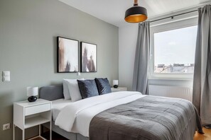 Un dormitorio moderno con una cama, dos mesitas de noche, un velador, un ventanal con cortinas y dos obras de arte colgadas en la pared.