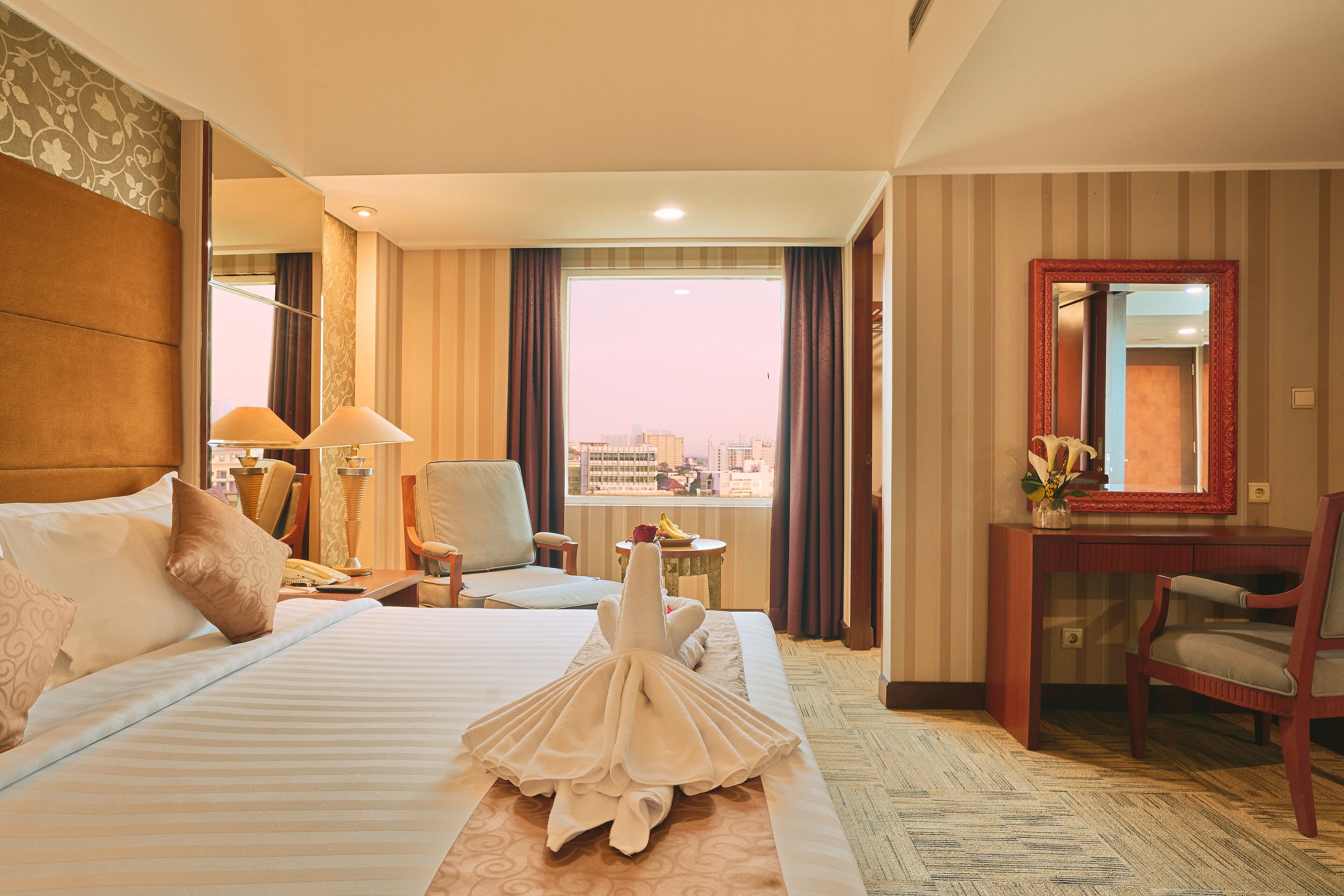 Habitación de hotel con una cama grande, un escritorio, una silla, una mesita con un florero y vistas a la ciudad por la ventana.