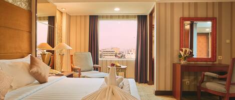 Habitación de hotel con una cama grande, un escritorio, una silla, una mesita con un florero y vistas a la ciudad por la ventana.