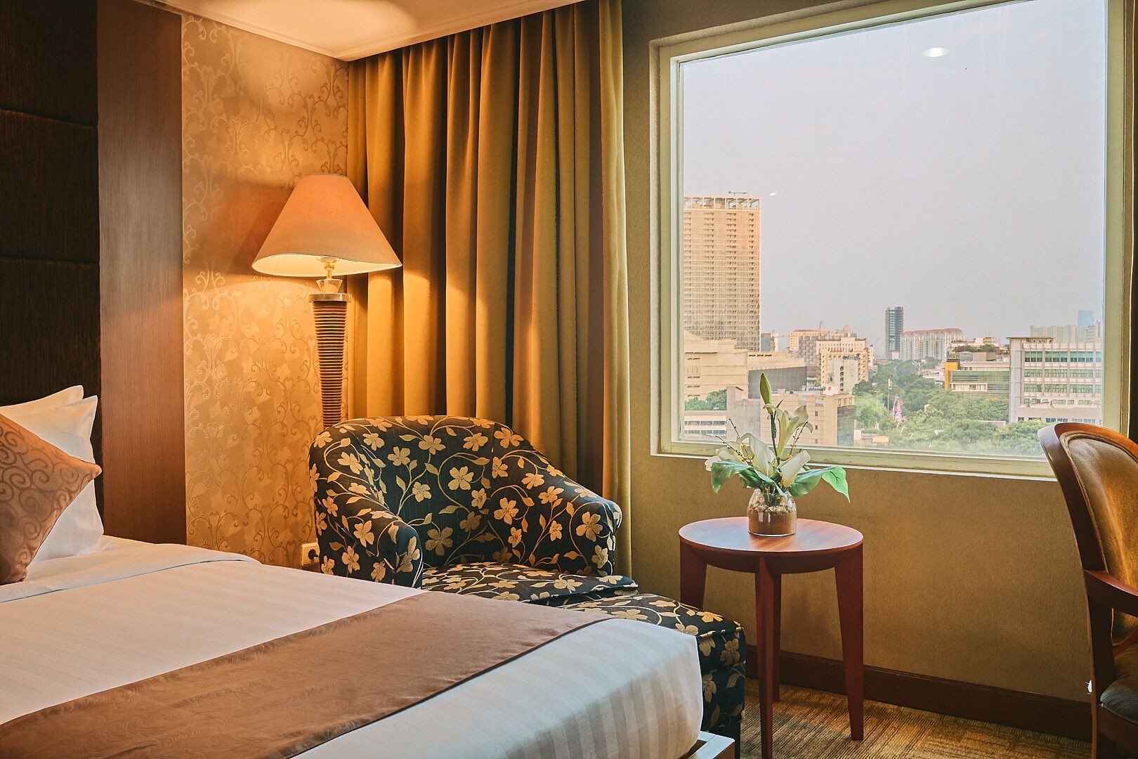 Habitación de hotel con una cama, un sillón floral, una mesita con una planta y vistas a la ciudad a través de una ventana.
