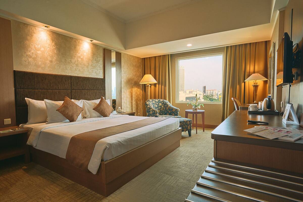A hotel room with a large bed, a desk with a chair, a window with curtains, and a city view.