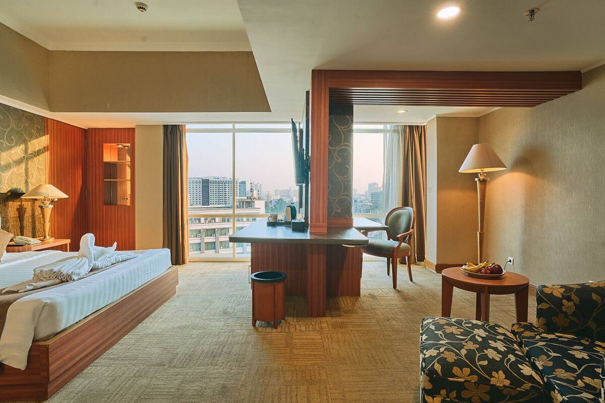 A hotel room with a bed, desk, chair, and a view of the city.