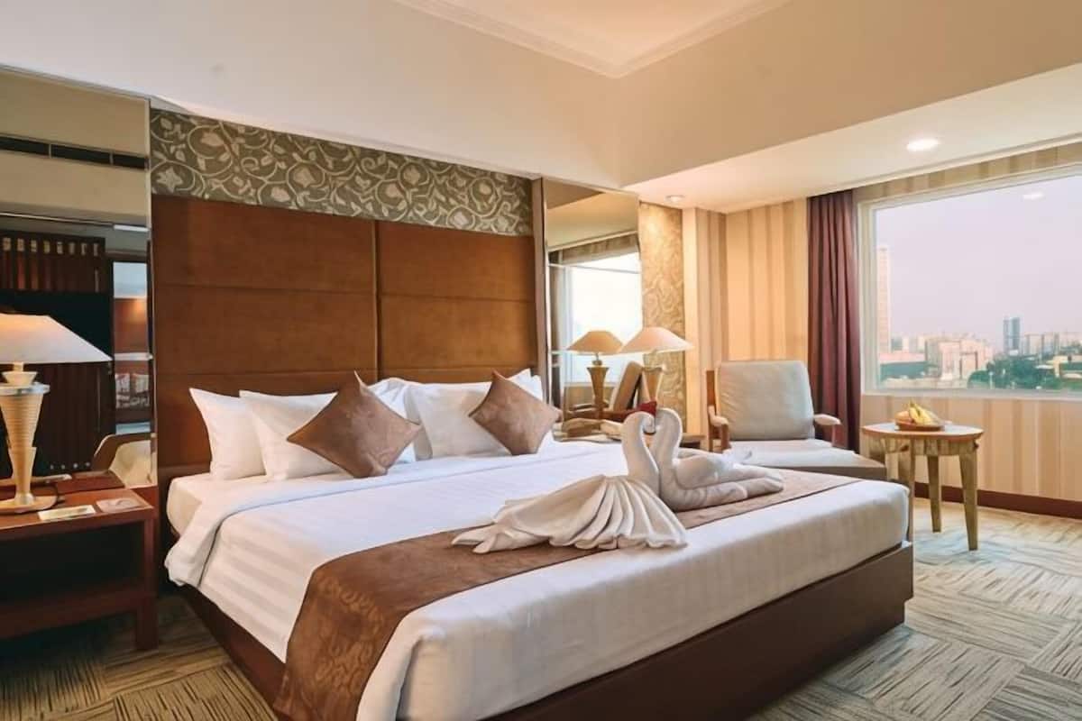 A hotel room with a large bed, a city view, a desk with a lamp, and a chair.