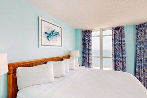 A hotel room with a large bed, a framed artwork of a blue bird, a window with curtains, and a view of the sea.