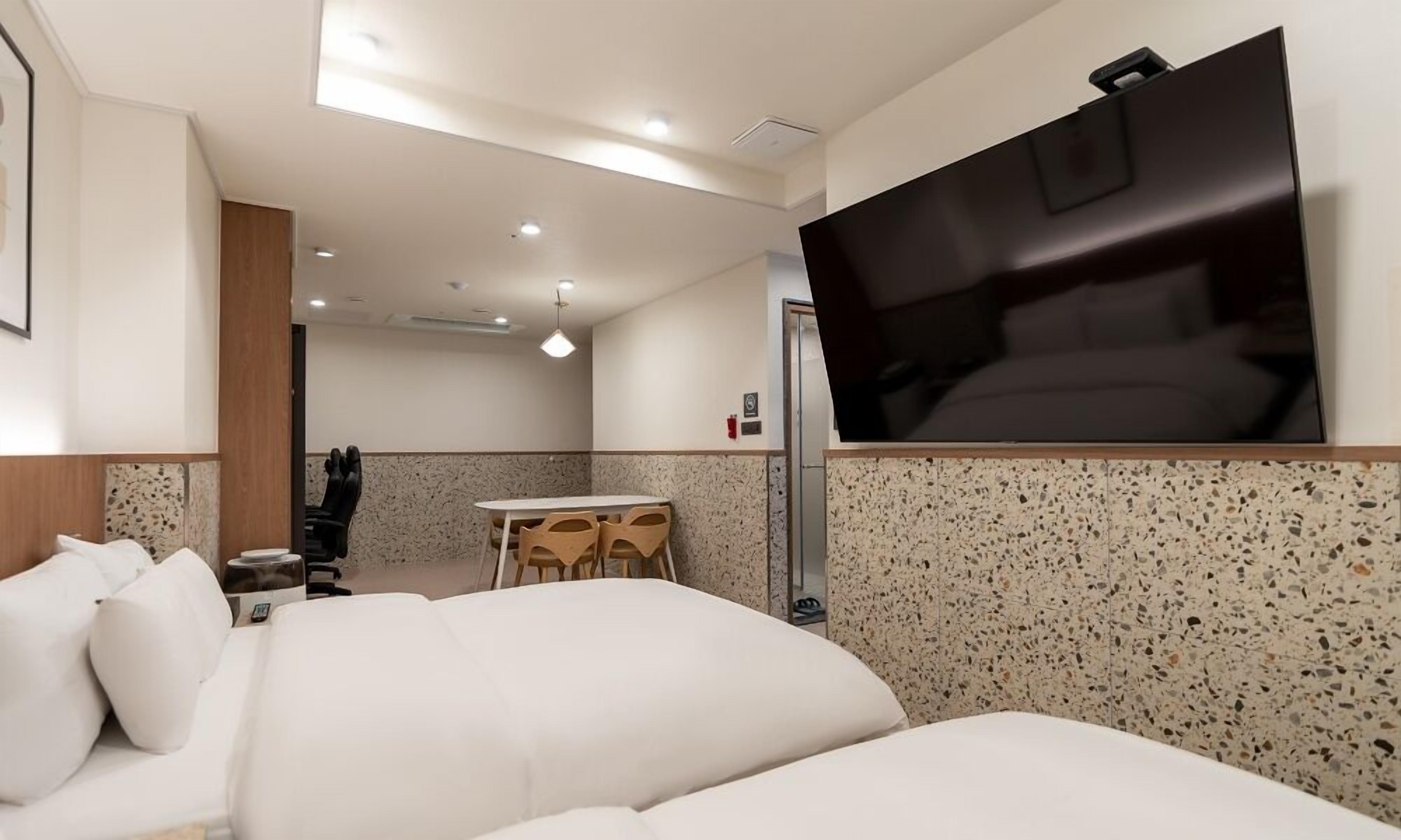 A modern hotel room with a large bed, a flat-screen TV mounted on the wall, a small dining table with chairs, and a kitchenette area.