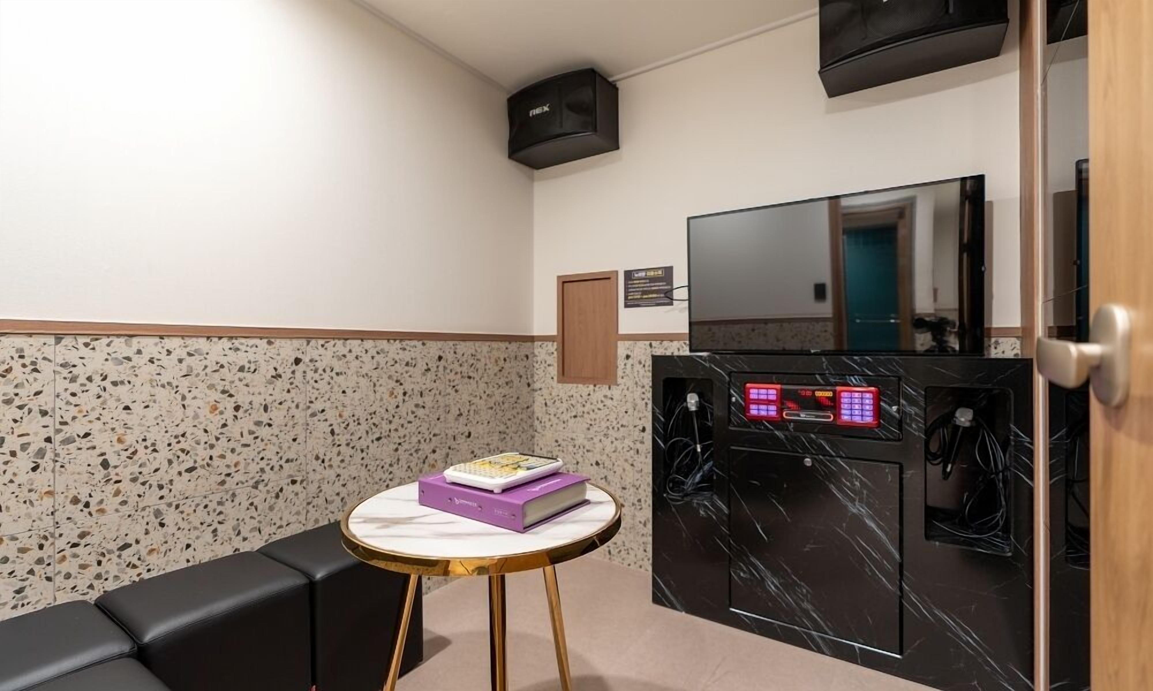 A hotel room with a black leather sofa, a small round table with a white marble top, a black marble fireplace, a flat-screen TV mounted above the fireplace, and a wooden door.