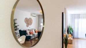 Ein modernes Wohnzimmer mit Sofa, Couchtisch und Sesseln, einem runden Spiegel an der Wand und einem großen Fenster mit Vorhängen.