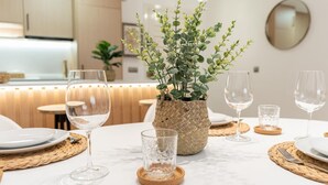 Ein Essbereich mit einem Tisch, gedeckt für vier Personen, mit gewebten Tischsets, klarem Glas und einem Korb mit Grünpflanzen.