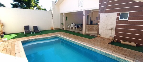 A swimming pool with a tiled deck, two lounge chairs, and a small patio area with a table and chairs.